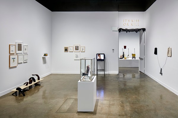 Installation view of a gallery featuring framed prints on the walls and small objects inside a glass container in the center of the room.&nbsp;