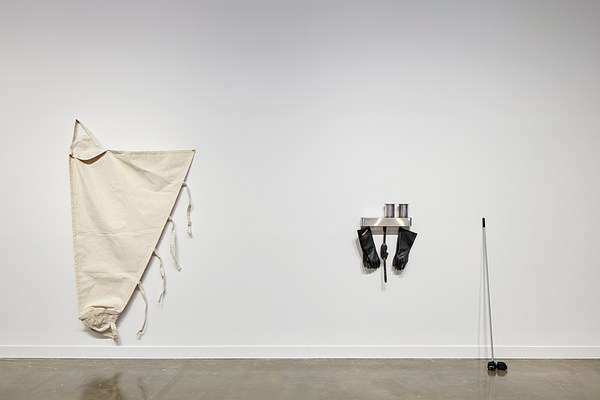 Cloth sculpture on the left with a triangular shape and a hanging sculpture on the right with a metal shelf, black rubber gloved, and a pole leaning against the wall.&nbsp;