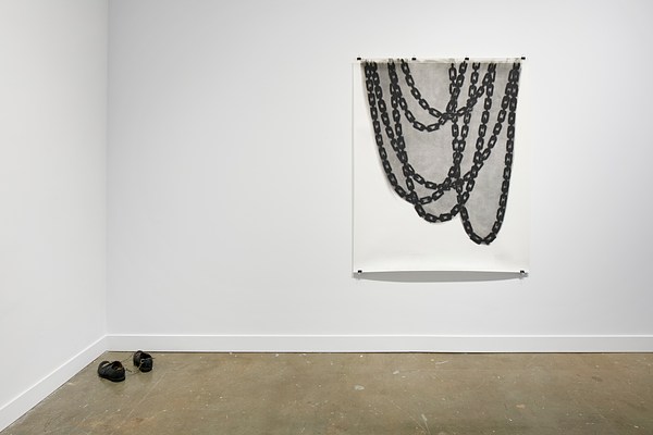 White tapestry with a grey and black painting of chain link on it and a pair of black shoe sin the left corner attached with a chain.&nbsp;