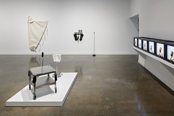 Installation view featuring two sculptures on the back wall, a metal cart on a white platform on the left and repeating screens on the right with an image of a shirtless man on them.&nbsp;