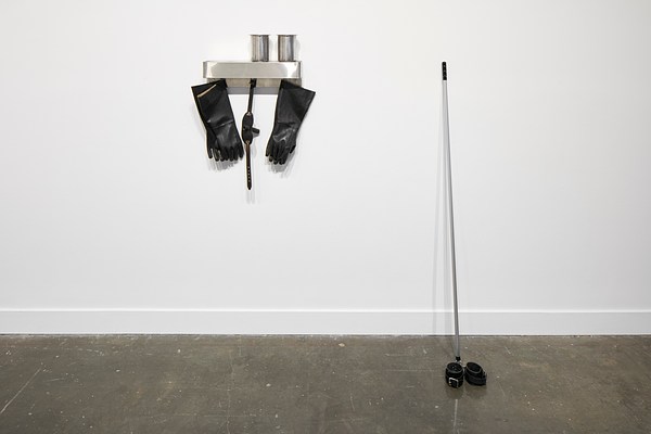Sculpture featuring a metal shelf with two cups on it and black rubber gloves hanging from it and a metal pole on the right with two small black belts wrapped in front of it.&nbsp;