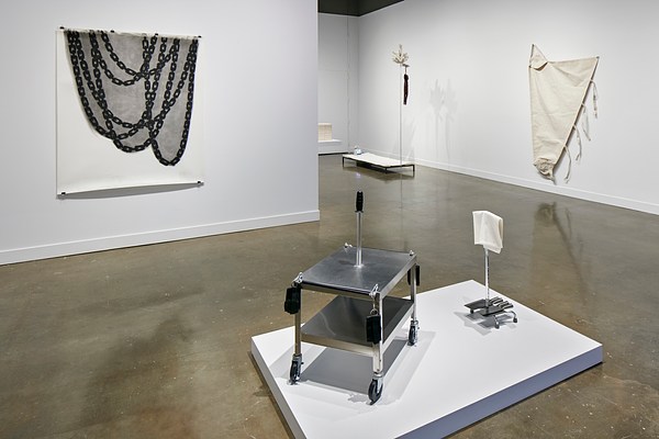 Tapestry on the left with a gray and black painting of chain link, a metal stand on a white platform in the center and two smaller sculptures in the background.&nbsp;