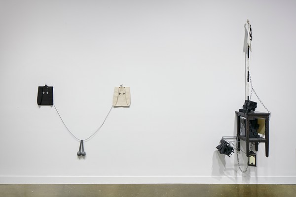 Installation view with two small bag like shapes on the left attached by a wire and two balls on a chain hanging from the center of the wire. On the right, there is an industrial looking sculpturing featuring a small side table and chains.&nbsp;