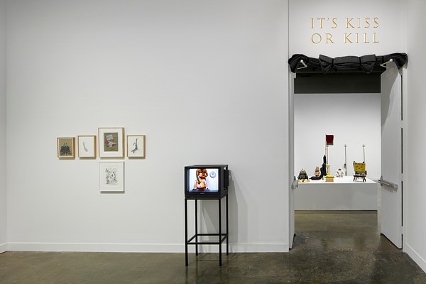 Five framed prints on the left next to a tv screen with a shirtless man sitting down and another man behind him with his hands on the mans chest and shoulder. On the right, a doorway leads into another gallery room with the title "It's Kiss or Kill" above the doorway.&nbsp;