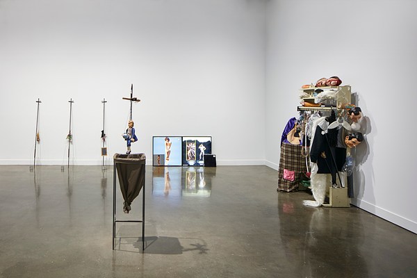 Installation view featuring a cluttered clothing shelf on the right with puppet heads and cloths and smaller sculptures on the right.&nbsp;