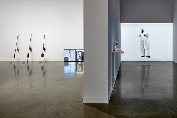 Large projector on the right with a man wearing a bunny costume. On the left, three small sculptures lean against the wall.&nbsp;