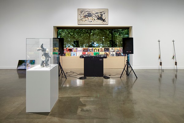 Installation view with a large window in the center and wooden shelves lined with records behind a speaker and dj booth. A glass cased bugs bunny sits on a white podium facing the window.&nbsp;