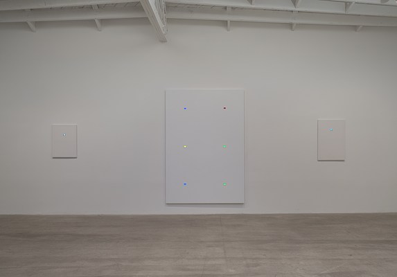 Installation view of a gallery space with a white wall and grey, concrete floor. On the white wall, we can see three paintings hung with equal space between them. They are all white, rectangular, in portrait format and have photographic slides in color mounted to them. The painting in the center is large-scale whereas the two on the sides are small-scale. In the ceiling, we can see parts of white beams in vertical and horizontal directions.