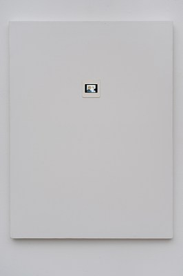Close view of a painting seen in previous installation images hanging on a white wall. The painting is in portrait format, and primed in white. Above the center of the painting, a photographic slide has been mounted directly onto the canvas. The photo slide appears to be of a painting. In the slide, we can see two people. To the left, we see the back of a person with brown shoulder length hair and a blue jacket with a white collar. To the right, we can see a man with light skin and brown short hair, wearing a grey shirt. He is looking at the person in blue jacket, and they appear to be engaged in a conversation. The slide is framed by a black frame, and it sits within a white paper frame. The slide appears lit from behind. Below the painting, we can see a subtle drop shadow on the wall.