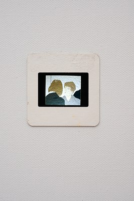 Close view of the photographic slide seen in previous image. The photo appears to be of a painting. In the slide, we can see two people. To the left, we see the back of a person with brown shoulder length hair and a dark jacket with a white collar. To the right, we can see a man with light skin and brown short hair, wearing a grey shirt. He has light blue eyes, and is looking at the person in dark jacket, and they appear to be engaged in a conversation. They are in a room with white walls, and in the background we can see what looks like the shoulder of someone wearing black. The photo is framed by a black frame, and it sits within a white paper frame. The slide appears lit from behind. Around the slide, we can see the grain of the canvas underneath the layer of white prime.