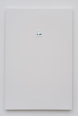 Close view of a painting seen in previous installation images hanging on a white wall. The painting is in portrait format, and primed in white. Above the center of the painting, a photographic slide has been mounted directly onto the canvas. The photo slide appears to be of a room with a white wall and grey floor. Against the wall, two paintings are leaned, and on the foreground on the floor, we can see some colorful objects of which one is box-shaped. The slide sits within a white frame, and appears lit from behind. Below the painting, we can see a subtle drop shadow on the wall.