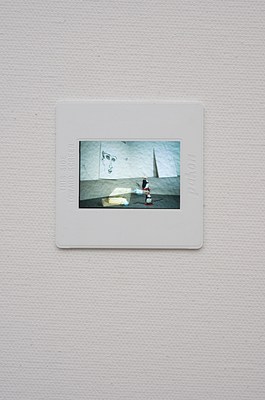 Close view of the photographic slide seen in previous image. We can see a white wall in the upper half of the photo, and a grey floor in the lower half. We can see two paintings leaned against the wall: to the left, a white primed canvas with a face outlined in black, and to the right, a white canvas with handwritten text in blue. On the floor, to the left, we can see a rectangular wooden box with a light blue cylinder-shaped object attached to its right side. To the right of the box, we can see a sculpture of a cartoon penguin that is connected to a power plug next to the painting of a face via a black cable. The lower half of the photo consists in a superimposition. The penguin sculpture and the box are doubled, and seen twice from two different angles. The slide appears lit from behind, and sits within a white frame with the text "pakon" vertically embossed on its right side. On the left side of the frame, we can read "This Side Toward Screen" written in caps. Around the slide, we can see the grain of the canvas underneath the layer of white prime.. . . 