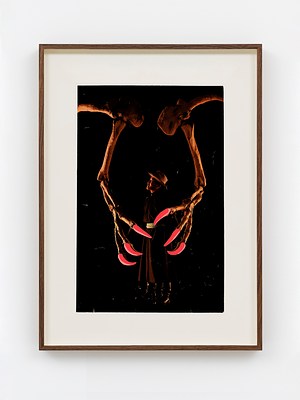 A framed, vertically oriented image of large hand or wing bones reaching down to clutch a person. The bones appear to be photographs but large red/pink nails or talons have been added at their ends. The pink nails appear cartoonish or photoshopped with a perfect smooth texture. The figure centered in the image also seems to be a photograph. The figure is a man with brown skin, wearing a hat, a long brown robe, and tall black boots. His face appears contemplative rather than afraid. The background of the picture is dark and the figure almost totally blends into the background.