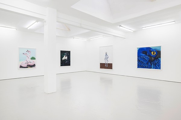 An installation view of a brightly lit gallery with white walls, ceiling and skylights and a pale, shiny floor. Four paintings are hung across the two back walls of the gallery. On our left, we can see a square painting of two pale pink dinosaurs; next, a mostly dark painting of a dinosaur with a white beak, and to the right, a pale gray standing dinosaur that resembles a human figure. The last painting is bright blue, and is a close up of the side of a dinosaur's head.