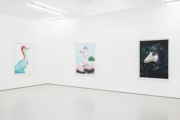 A tighter installation view of a brightly lit gallery space. The view shows the corner of the gallery. On our left, we can see a vertically oriented painting of a pale turquoise dinosaur with an orange mohawk of feathers. On the other wall, to our right, two pale pink dinosaurs sit together, their long necks entwined. The last painting is mostly dark green and black. The green of leaves melds into the dark feathers of the dinosaur. A bright white beak is centered in the painting.