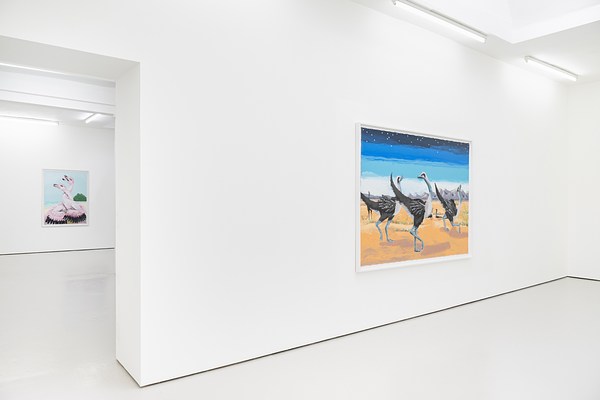 An installation view of a white gallery with a doorway to our left that leads to another room in the gallery. On our right, we can see a large painting of three ostrich like birds. On our left, through the doorway and in the second room, a pale painting of entwined birds hangs on the back wall.