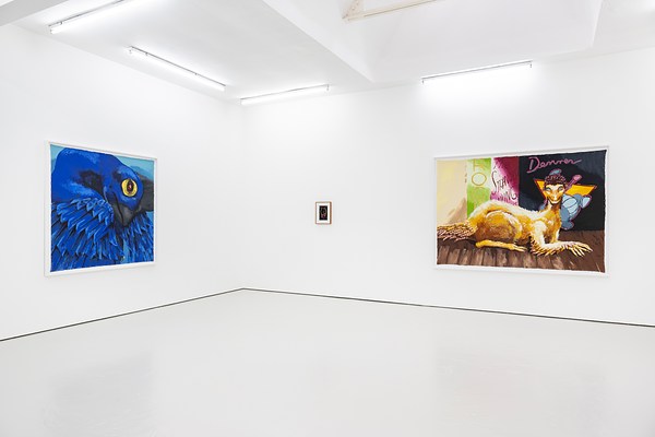 An installation view of the corner of a gallery. On the left wall is a large, cobalt blue painting. A yellow eye glows out of the close up of a dinosaur's face. On the wall to our right is a small framed photograph or drawing. It's dark and too small to see, but after zooming, it appears to be the arms or wing structure of a bat or dinosaur. To it's right is a large horizontally hung painting of a yellow dinosaur. It is lying on the floor like a cat or a sphinx and behind it colorful banners have words like "Rock" and "The Strangest Thing."