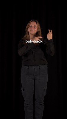Another dark, vertically-oriented image of the woman in black signing at the camera. The light skin tone of her hands, face, and hair stands out in contrast to the dark background. Her right forearm crosses her chest, and she holds her right index and middle fingers together, pointing at her left hand. She is holding her left hand upright, index and middle finger extended in a kind of backwards peace sign, with the back of her hand facing the camera. Over her chest is the caption "look-back" in white, sans serif font.