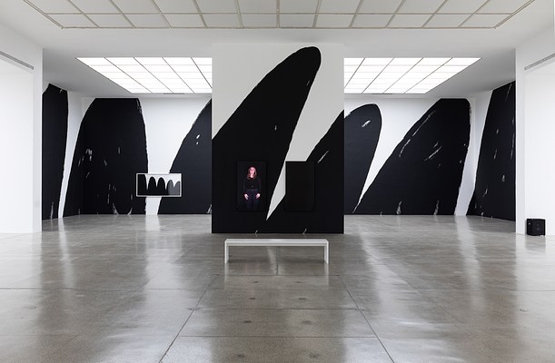 Tighter installation view from the main space towards the front section of the gallery, showing the black and white graphics covering the walls of that section. The image is symmetrical. On the side of the freestanding wall facing us are two video monitors hung vertically, with a white bench positioned in front of them. The one on the left shows a standing figure from the knees up wearing a dark top and pants. The one on the right is black. A framed artwork hangs on the left side of the wall behind the freestanding wall. It looks like a smaller version of the black and white graphics of peaks and valleys covering the walls on which it's hung.