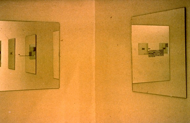 Installation view of a corner in a room and works on the walls. Two walls meet in the center of the image. On our left, we can see a work in the form of a mirror with small, black text in the center, hanging on the wall. In the mirror, we can see the reflection of the right wall from out of frame, and three works in the form of mirrors with text in the center of the same square dimensions. On the right wall, we can see another mirror with black text in the center. Reflected in this mirror, we can see another mirror of the same series, hanging on the left wall out of frame. In that mirror, we can see two additional mirrors reflected from elsewhere in the room. The image is in greyscale with a tint of orange.