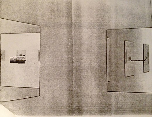 Tighter installation view of a corner in a room and works on the walls. Two walls meet in the center of the image. On the wall to our left, we can see a work in the form of a mirror with small, black text in the center, hanging on the wall. In the mirror, we can see the reflection of the right wall, out of frame, and a work in the form of a mirror with text in of the same series. In the mirror, we can see three additional mirrors of the same series reflected from elsewhere in the room. On the right wall, we can see another mirror with black text in the center. Reflected in this mirror, we can see three mirror works from the same series hanging on the left wall out of frame. The image is in greyscale and has the texture of a photo copy.