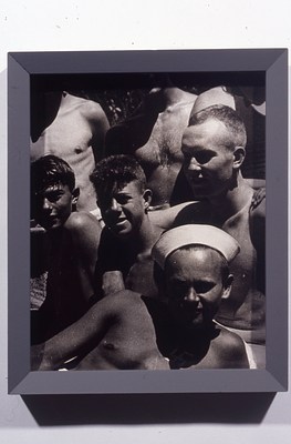 Black and white photo in a gray frame cropped from a larger group photo of young men with light to medium-light skin tones and short hair, many of whom are shirtless. In this cropped image, the reclining figure in what looks like a sailor's cap is in the foreground and takes up the bottom third of the image.  On closer inspection, in the shadow cast by his face onto his chest, it's possible to make out a white swastika. Behind him is a row of three young men, two with their arms draped over each other's shoulders. Behind this row is another row of three bare torsos.