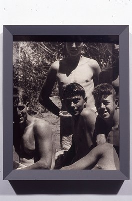 Black and white photo in a gray frame, showing a small group of shirtless young men with light to medium-light skin tones and short hair, all smiling at the camera. Some of the figures are only partially in frame, indicating that this image was cropped from a larger photograph. They are outdoors in direct sunlight. The figure on the bottom left has tan lines from a tank top.