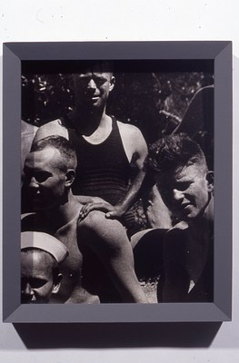 Black and white photo in a gray frame cropped from a larger group photo of young men with light to medium-light skin tones and short hair, many of whom are shirtless. At the center of this image, a hand rests on a bare, muscular shoulder between two figures who look directly at the camera, squinting in the sun. At the bottom left is a figure wearing what looks like a white sailor's cap.