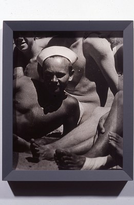 Black and white photo in a gray frame cropped from a larger group photo of young men with light to medium-light skin tones and short hair, many of whom are shirtless. In this cropped image, a reclining, shirtless young man wearing what looks like a sailor's cap squints at the camera. On closer inspection, in the shadow cast by his face onto his chest, it's possible to make out a white swastika. Parts of other figures crowd around him — for instance, a bare torso behind him, and two pairs of feet and shins, which rest in front of him on the right side of the image.