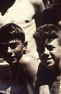 An even closer crop of the photo with two figures squinting at the camera with other figures crowded around, partially in frame. The figure on the left appears to have damp hair.