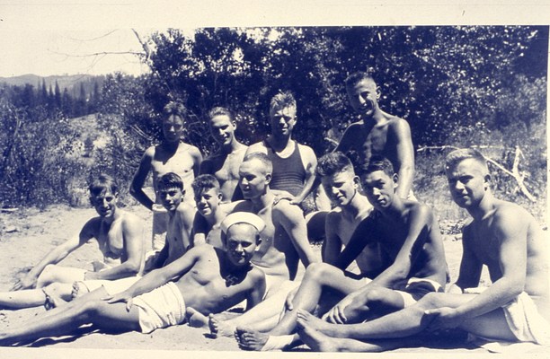 The full group photo of young men with light to medium-light skin tones and short hair, many of whom are shirtless. There are twelve figures sitting, kneeling, or reclining together on a sandy beach in front of some shrubby foliage. Some appear to be nude except for a towel draped over or wrapped around their waist. In this image, the swastika on the chest of the central figure is especially pronounced.