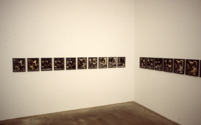 Installation view of the two rows of framed pictures on each of two walls forming the corner of the gallery. The subject matter is difficult to make out, but there appears to be some repetition from picture to picture.