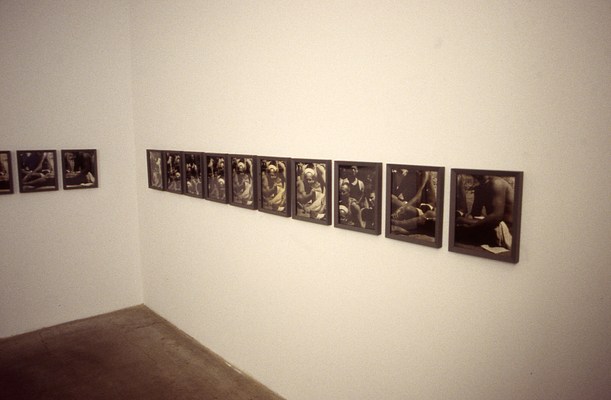 Installation view of the two rows of framed pictures on each of two walls forming the corner of the gallery, with mainly the row on our right visible. The pictures appear to be framed, black and white photographs, each one cropped from a larger photo of a group of muscular figures, many of whom are shirtless. One reclining figure at the front of the group is wearing what looks like a sailor's cap.
