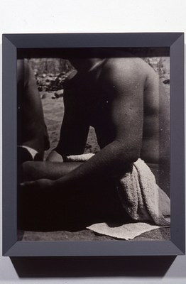 Black and white photo in a gray frame cropped from a larger group photo of young men with light to medium-light skin tones and short hair, many of whom are shirtless. This image is cropped to show only a seated figure from knee to shoulder, bare except for a white towel draped over his lap, and the back of another figure seated beside him on the sand.