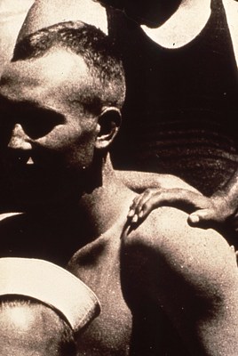 An even closer crop of the image of a hand resting on a bare, muscular shoulder between two figures, the one at the bottom left wearing white sailor's cap.