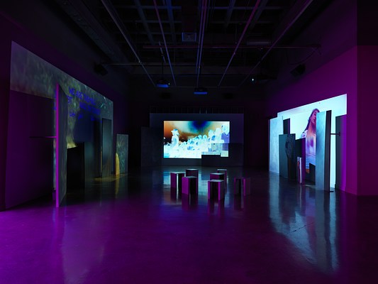 Videos are projected onto uneven surfaces of large panels against three walls. The central projection shows glowing, almost psychedelic inverted footage of a group of figures in a line. The right projection shows a woman dancing, and the left projection shows a person wearing a yellow shirt overlaid with text in English and Arabic, reading “We are tremble / In debt / And in doubt.”