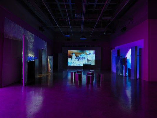 Videos are projected onto uneven surfaces of large panels against three walls. The central projection shows glowing, almost psychedelic inverted footage of a group of figures in a line. The right projection shows a woman (also in unnatural, inverted tones) dancing, and the left projection shows a person wearing a yellow shirt. All three images are overlaid with text in English and Arabic reading, “Unbound.”