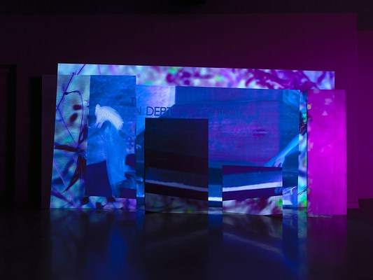A saturated blue and purple video is projected onto an uneven surface of overlapping rectangular panels. The rightmost panel is illuminated pink, while the rest of the projection is tinted deep blue. Onscreen, abstracted video footage of plants and a person’s arm coming into the frame on the right is overlaid with text in Arabic and English that reads “In debt.”
