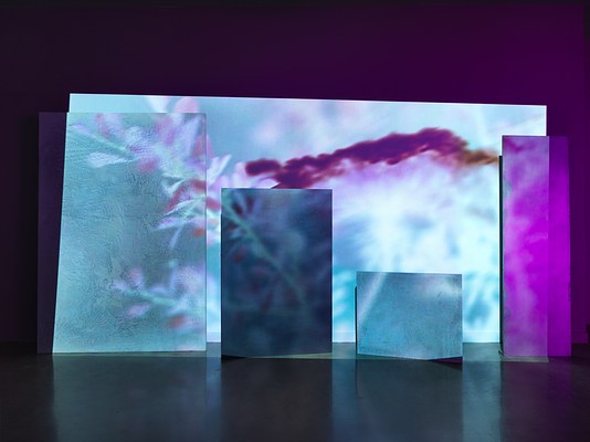 A saturated cyan and purple video is projected onto an uneven surface of overlapping rectangular panels. Onscreen is a close-up footage of leaves and flowers in surreal tones.