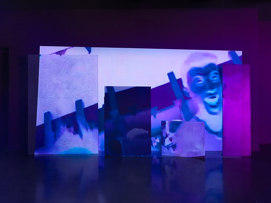 A saturated blue and purple video is projected onto an uneven surface of overlapping rectangular panels.The rightmost panel is illuminated pink, while the rest of the projection is tinted deep blue. Onscreen, a person’s face with their mouth open is shown in inverted tones.