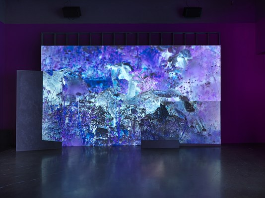 A saturated blue and purple video is projected onto an uneven surface of overlapping rectangular panels. Abstracted, high-contrast, video footage of plants fills the frame of the video.