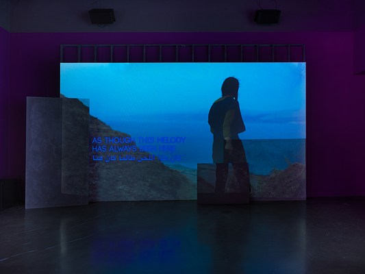 A deep blue video depicting a person looking out from a mountain onto a body of water is projected onto an uneven surface of overlapping rectangular panels. Overlaid on the video footage, English and Arabic text reads “As though this melody has always been here”.