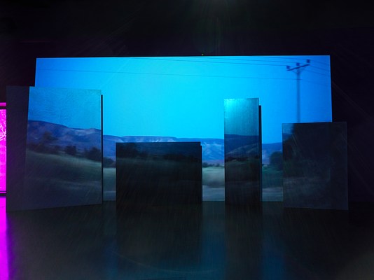 A blue and green depicting a landscape is projected onto an uneven surface of overlapping rectangular panels. Onscreen, an electrical pole hangs in the upper right corner with wires stretching horizontally across the frame. Mountains fill the background. Left of the projection, a small sliver of purple light peers through a window into the room.