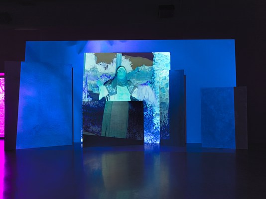 A saturated blue video is projected onto an uneven surface of overlapping rectangular panels. Onscreen, an abstracted woman wearing a hijab and glasses has her arms outstretched facing the camera with a landscape of various plants behind her. Left of the video projection, a small sliver of purple light peers through a window into the room.