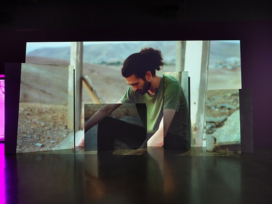 Video footage of a man wearing a green shirt sits in a dry landscape looking toward the ground surrounded by rocks and vertical planks. The video is projected onto an uneven surface of overlapping rectangular panels. Left of the video projection, a small sliver of purple light peers through a window into the room.