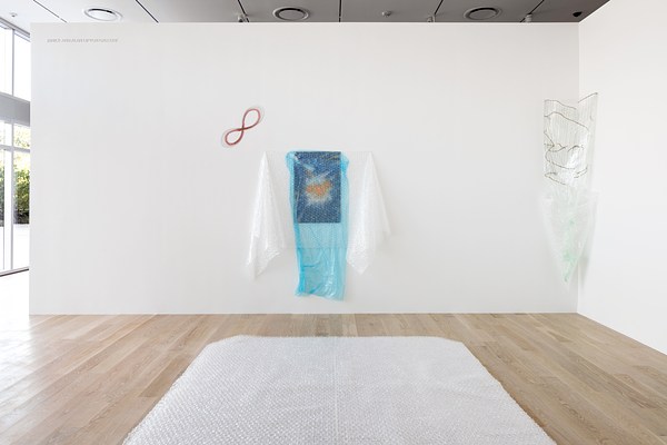 An installation view of a brightly lit gallery space. Two white walls make a corner on our right and a wall of windows extends to our left beyond the frame of the photo. A large sheet of clear bubble wrap, or a stack of sheets, is laid on out the floor like a rug. On the wall behind the stack of bubble wrap is an abstract drawing with some bright orange shapes at center, veiled by a sheet of semi-transparent, blue plastic tacked up in front of it, somewhat messily. Hung horizontally over these two layers is a sheet of clear bubble wrap, pinned at the top so that the excess material hangs down loosely, as would towels on hooks. Above and to the left of this piece is a diagonal figure eight or infinity symbol, drawn directly on the wall in what looks like red pencil and graphite. Installed in the corner on our right is a piece made of wire, plastic, and bubblewrap.