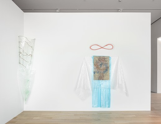 Installation view of the gallery with the corner piece made from plastic, wire, and bubble wrap on our left and another piece with garment proportions on our right. In this piece, another abstract drawing or painting is veiled by a semi-transparent, light blue sheet of plastic, and over that, a sheet of clear bubble wrap pinned at the top so that the excess material drapes down like sleeves or towels on hooks. A horizontally-oriented, red infinity symbol drawn directly on the wall hovers, centered, above this assemblage.