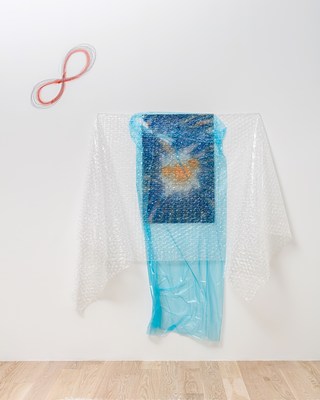 A closer view of the work with blue plastic and clear bubble wrap. An abstract drawing or painting with orange shapes at center in a field of navy blue is veiled by a sheet of semi-transparent, light blue plastic tacked up in front of it, somewhat messily. The blue plastic extends beyond the painting vertically, nearly reaching the floor. Hung horizontally over these two layers is a sheet of clear bubble wrap, pinned at the top so that the excess material hangs down loosely. If the piece were a garment, the place where the bubble wrap is tacked would correspond to its shoulders, and the excess lengths falling in folds would form sleeves. Floating above and to the left of the work is a diagonal figure eight or infinity symbol, drawn directly on the wall in what looks like red pencil and graphite.