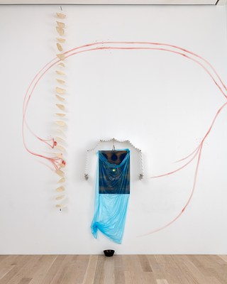 An installation view of a third wall work. A sheet of semi-transparent, light blue plastic wraps around another abstract drawing or painting of hazy dark blue and yellow circles on a brown background. The plastic drapes down at the top to reveal a bit of the drawing or painting's surface. The work is framed at the top by a paper accordion garland whose two ends form star shapes. To its left, a chain or mobile of what appear to be used coffee filters hangs down from the ceiling. Loose strokes of red paint or pencil drawn directly on the wall encircle the assemblage. A black bowl rests on the floor beneath it.