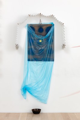 A closer view of the third wall work: a sheet of semi-transparent, light blue plastic wrapped around an abstract drawing or painting of hazy dark blue and yellow circles on a brown background. The plastic drapes down at the top to reveal a bit of the drawing or painting's surface. The work is framed at the top by a paper accordion garland whose two ends form star shapes. Towards the bottom, on our left, the bag has been tacked up to the wall. A black bowl rests on the floor beneath it.